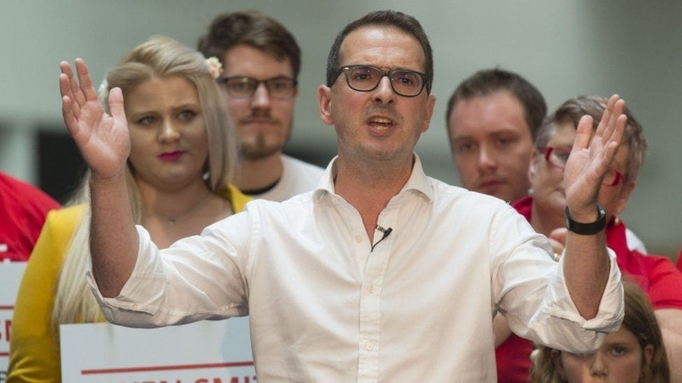 A white man in a white shirt, sleeves rolled up, wearing glasses and clip mic, gesturing with hands.